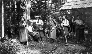 Six women in early 20th century hiking clothes, hats, and boots, hold their hiking sticks outside a wooden cabin in the woods